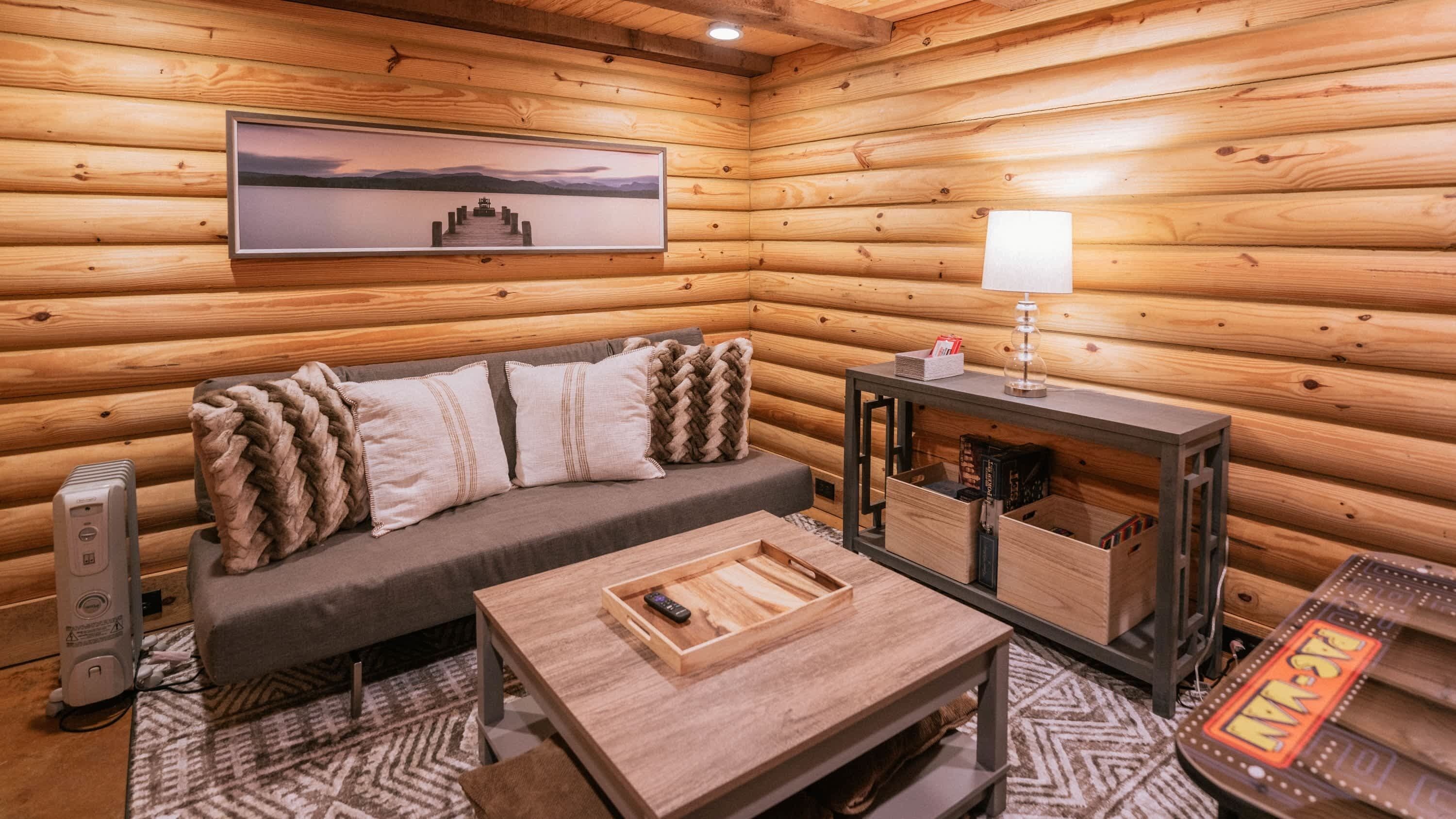 A cozy cabin interior with a wooden log wall, a sofa with cushions, a coffee table, a lamp, and a framed picture on the wall.