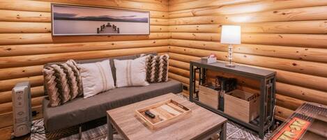 A cozy cabin interior with a wooden log wall, a sofa with cushions, a coffee table, a lamp, and a framed picture on the wall.