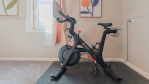 A stationary bike in a room with a carpeted floor, a window with curtains, and a wall with framed artwork. - Sloans Lake!  Walk to eateries/bars! Free parking! (Denver)