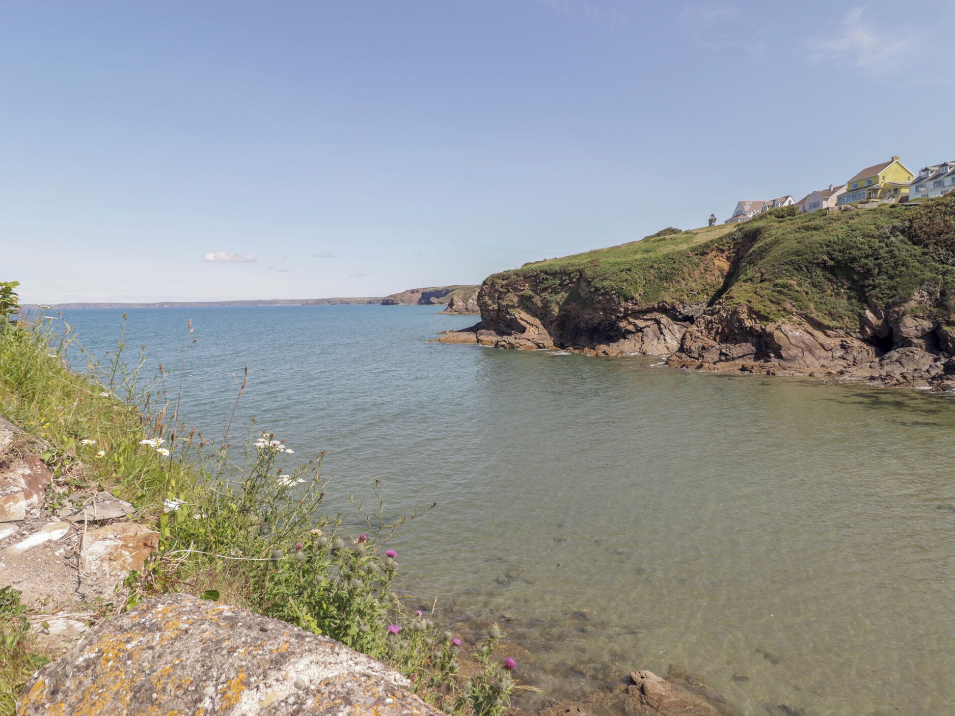 A coastal landscape with clear water, rocky cliffs, and a few houses on the hillside.