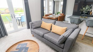A modern living room with a grey sofa, two yellow cushions, a wooden coffee table with brochures, and a dining area with a table set for four. - Driftwood Lodge (Haverfordwest)
