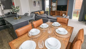 A modern kitchen with a built-in stove, a dining area with a wooden table set for four, and a living area with a sofa and TV. - Driftwood Lodge (Haverfordwest)