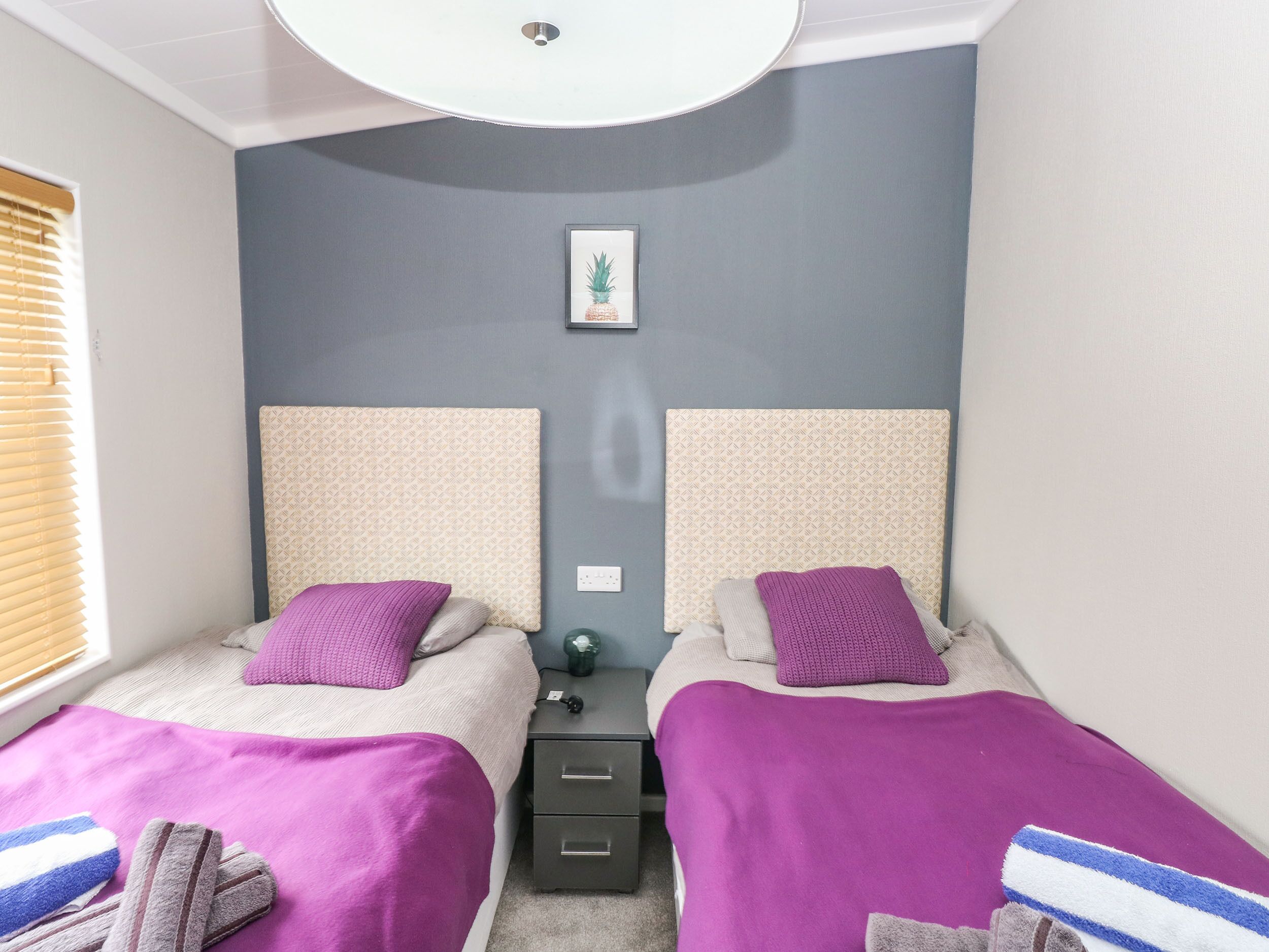 A bedroom with two beds, each with a headboard, a bedside table, and a nightstand. There is a framed picture on the wall above the beds.