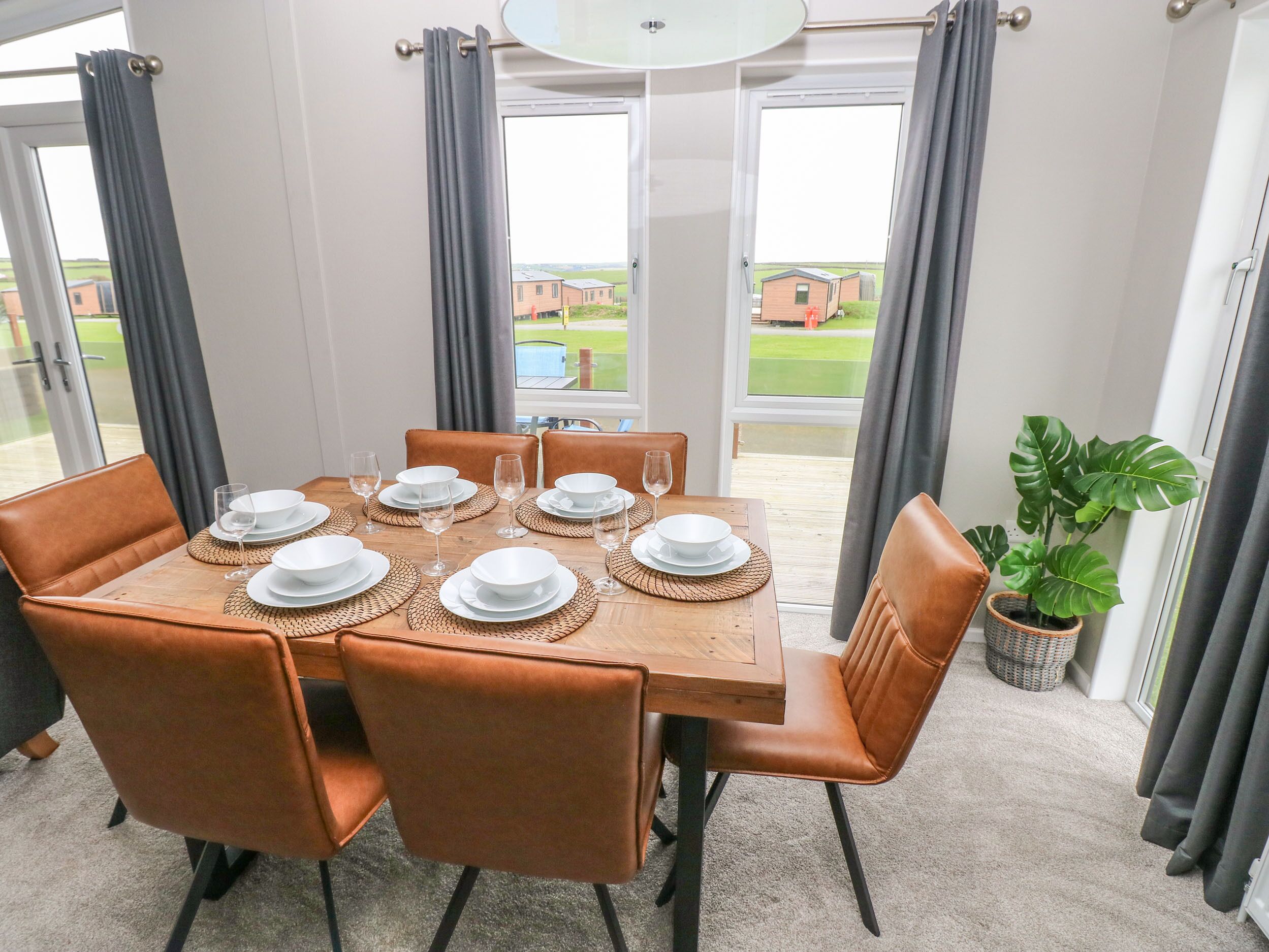 Un coin repas avec une table dressée pour six personnes, entourée de chaises marron, et offrant une vue sur un paysage verdoyant grâce à de grandes fenêtres.