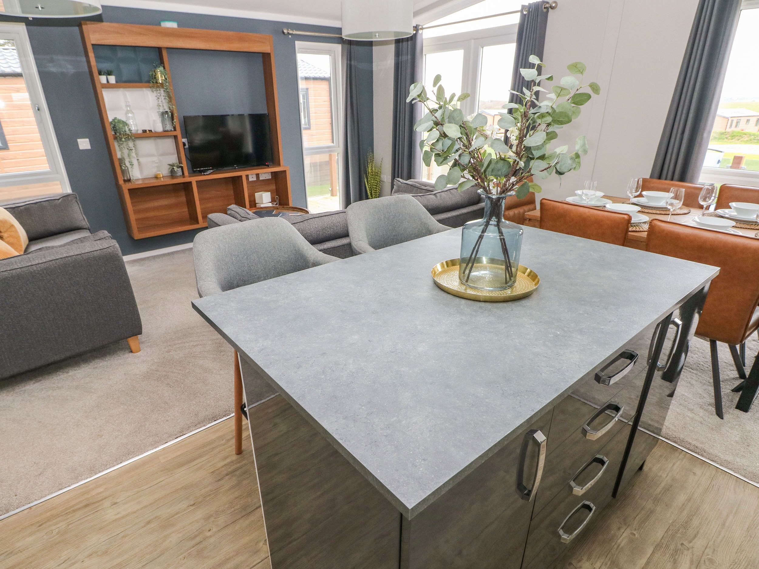 A modern living room with a grey sofa, a TV on a wooden stand, a dining table with chairs, and a vase with plants.