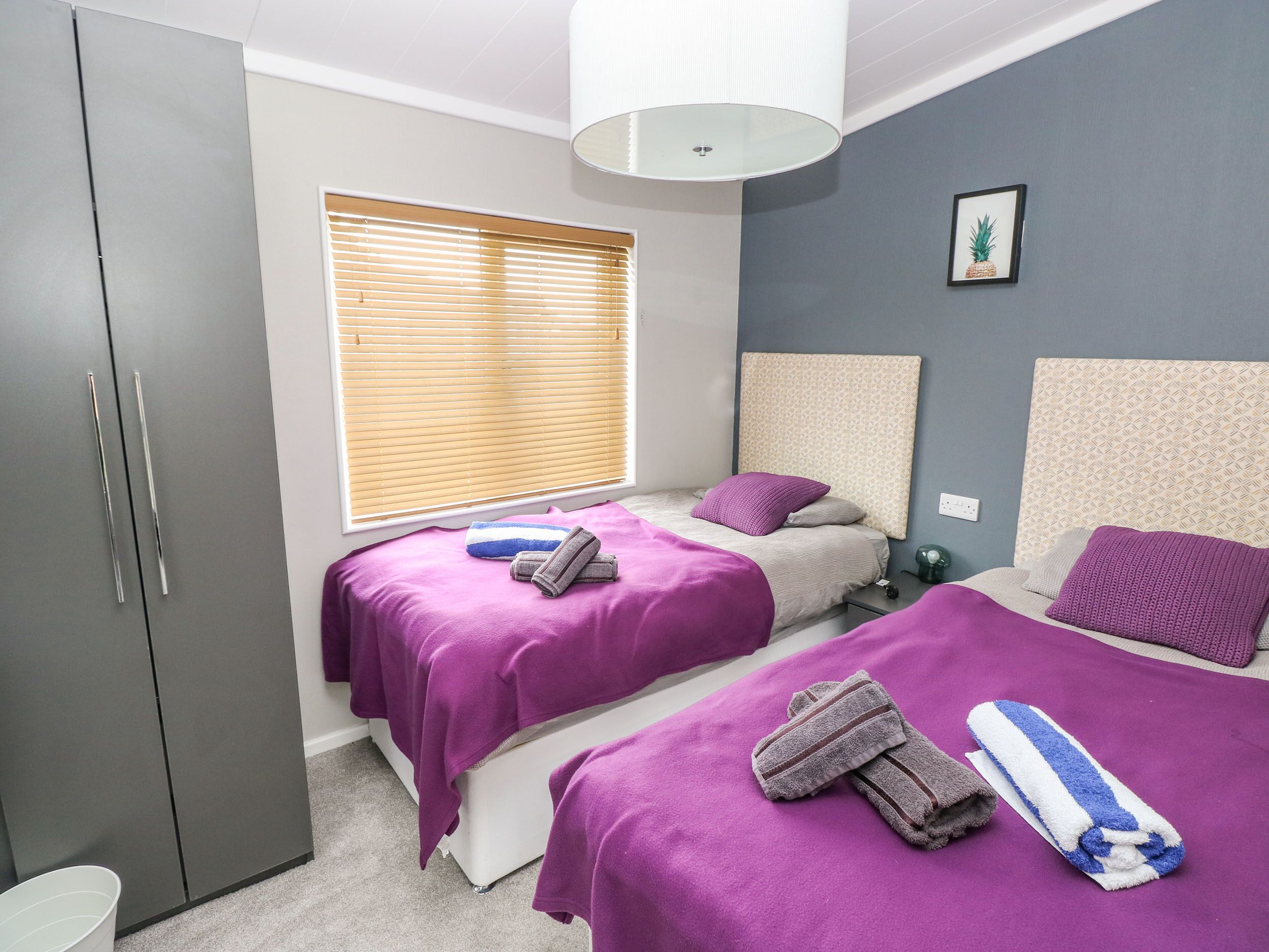 A bedroom with two beds, a wardrobe, a window with blinds, and a wall-mounted light fixture.