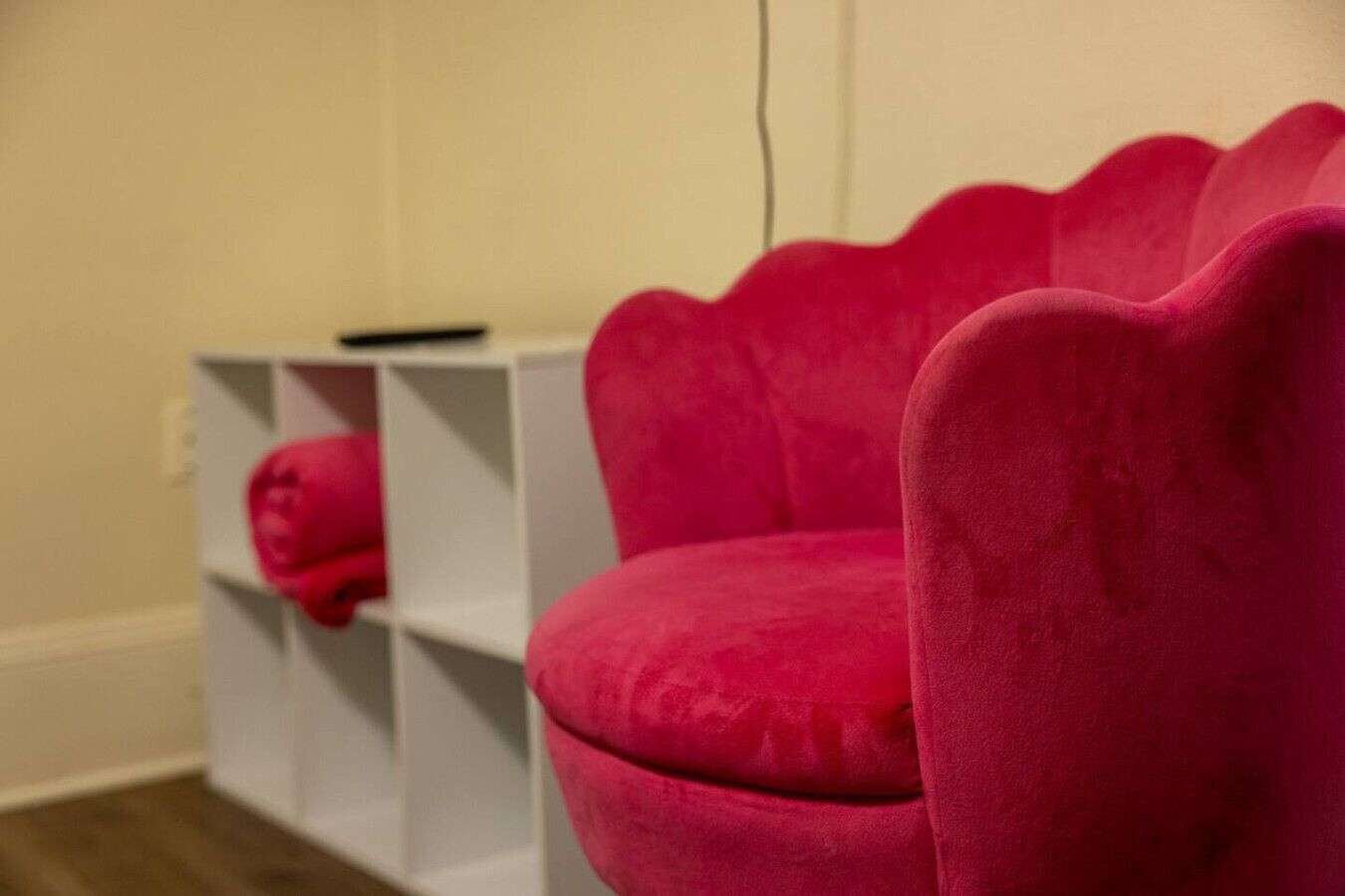 A pink chair with a scalloped design, a white shelving unit with a pink object on it, and a plain wall.