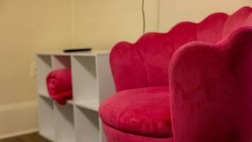 A pink chair with a scalloped design, a white shelving unit with a pink object on it, and a plain wall.