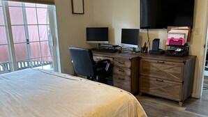 Un dormitorio con una cama, un escritorio con dos monitores, una silla, un aparador y un televisor en la pared.