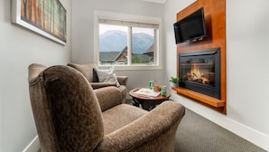 A cozy living room with a fireplace, a television mounted on the wall, and a large window offering a mountain view.