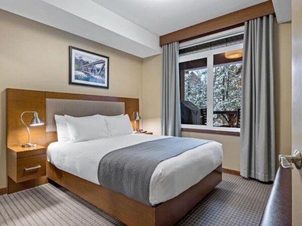 Habitación de hotel con una cama grande, una mesita de noche con lámpara, una ventana con cortinas y un cuadro en la pared.