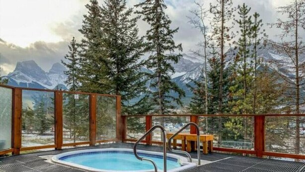 Un jacuzzi con vistas a montañas nevadas, rodeado de altos árboles y una terraza de madera.