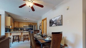 A dining area with a table set for four, surrounded by wicker chairs, a ceiling fan, and a kitchen in the background.