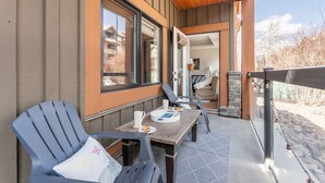 A wooden deck with two chairs and a table, a stone fireplace, and a view of a snowy landscape. - AURORA NIGHTS -  COZY LIVING -  SPA, POOL & HOT TUB (Canmore)