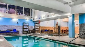 A swimming pool area with a blue and white color scheme, a seating area with blue cushions, and a staircase leading to an upper level. - AURORA NIGHTS -  COZY LIVING -  SPA, POOL & HOT TUB (Canmore)