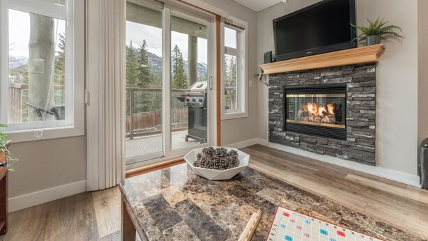 A modern living room with a stone fireplace, a flat-screen TV, and a large sliding glass door leading to a deck with a grill. - BLUE BIRD SKIES - MOUNTAIN VIEWS - GYM & SECURE PARKING (Canmore)