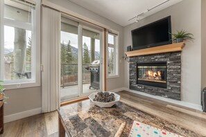 Una sala de estar moderna con chimenea de piedra, televisor de pantalla plana y un amplio ventanal corredero que da a una terraza con una parrilla.