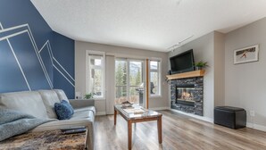 A modern living room with a stone fireplace, a sofa, a coffee table, and a TV mounted on the wall. - BLUE BIRD SKIES - MOUNTAIN VIEWS - GYM & SECURE PARKING (Canmore)