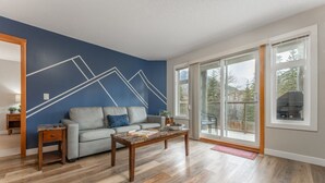 A living room with a blue accent wall, a grey sofa, a wooden coffee table, and large windows offering a view of the outdoors. - BLUE BIRD SKIES - MOUNTAIN VIEWS - GYM & SECURE PARKING (Canmore)
