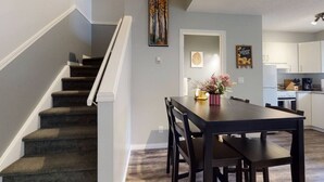 Una cocina moderna con armarios blancos, un comedor con mesa y sillas de madera oscura, y una escalera con peldaños cubiertos de alfombra oscura.