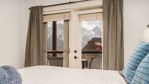 Ein Hotelzimmer mit Bett, zwei mit Vorhängen versehenen Fenstern, Bergblick und einem Balkon mit Stuhl.