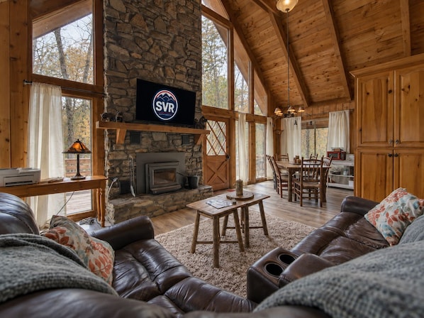 A cozy cabin interior with a stone fireplace, wooden furniture, and large windows.
