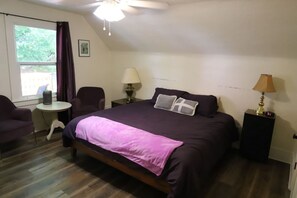 Un dormitorio con una cama, dos mesitas de noche, una silla, un ventanal y un ventilador de techo.