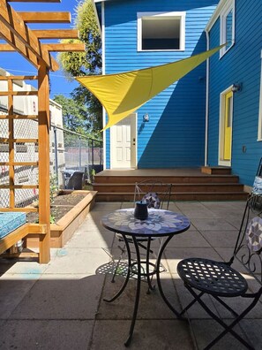 Un patio con una mesa y sillas, una pérgola de madera y una casa azul con una puerta amarilla.