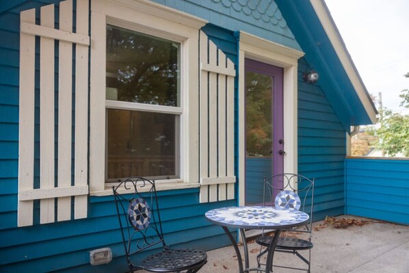 Una casa con fachada azul, una ventana blanca con persianas verticales, una puerta morada y un pequeño patio con dos sillas y una mesa de metal.