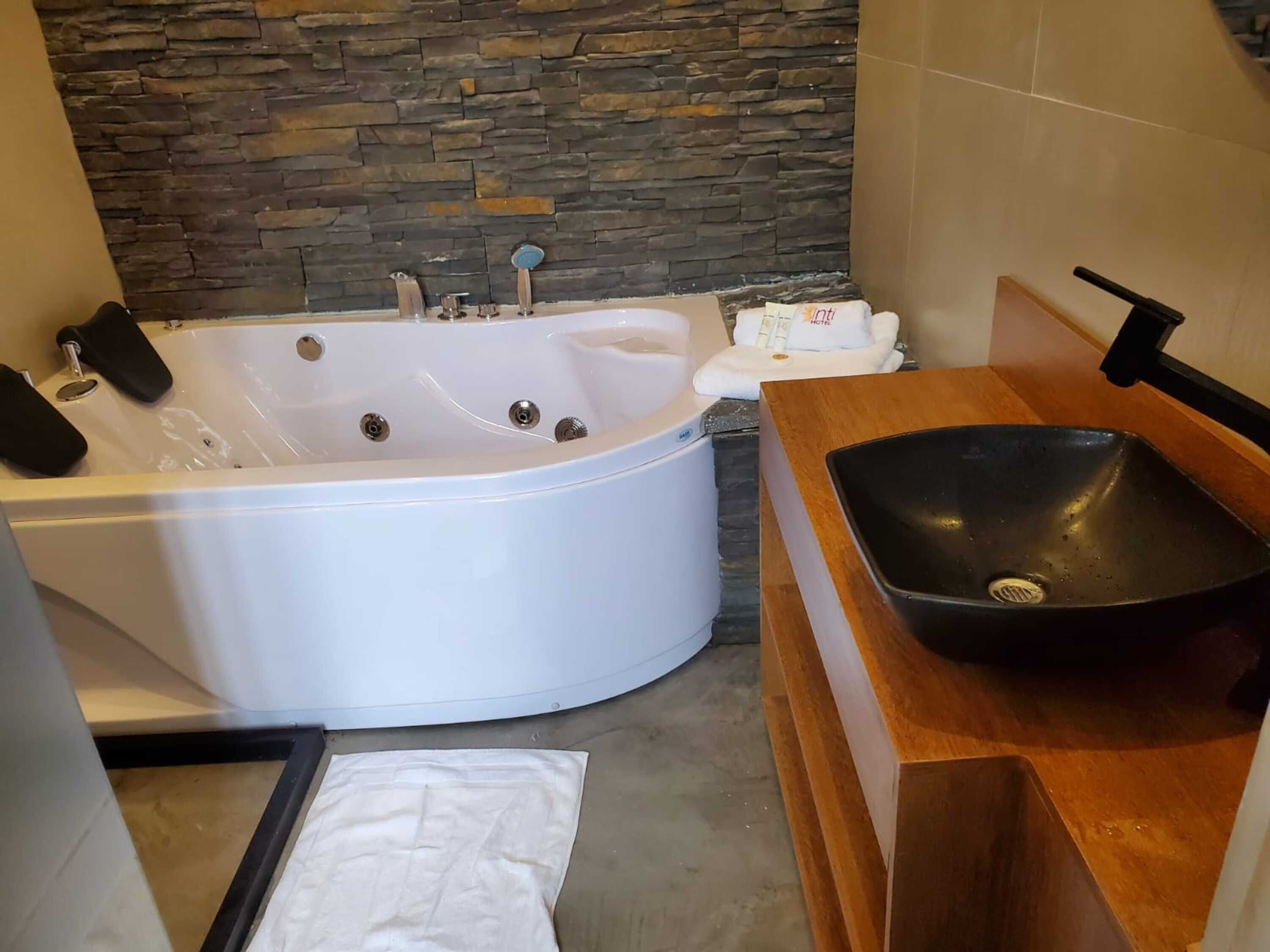 A modern bathroom with a stone wall, a white bathtub, a wooden vanity with a black sink, and a towel rack.