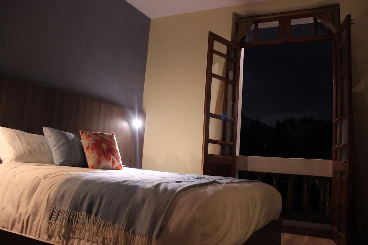 A bedroom with a bed, pillows, a nightstand, and a window with a view of the outdoors.