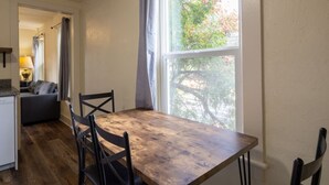 Een eethoek met een houten tafel, vier stoelen, een raam met uitzicht op bomen, en een keuken op de achtergrond.