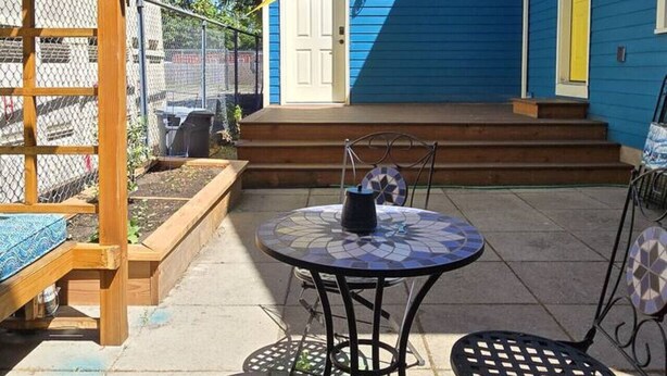 Een terras met een tafel en stoelen, een houten pergola en een blauw huis met een gele deur.