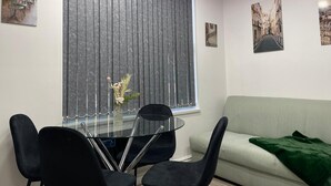 A modern living room with a glass dining table, black chairs, a grey sofa, and a green throw blanket.