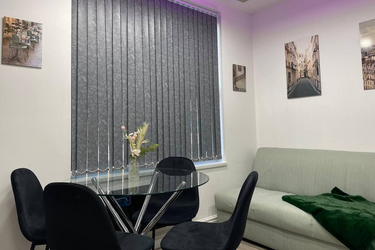 A modern living room with a glass dining table, black chairs, a grey sofa, and a green throw blanket.