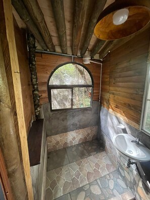 Um banheiro com piso de ladrilho de pedra, um espelho sobre bancada de madeira e uma janela com vista para vegetação.