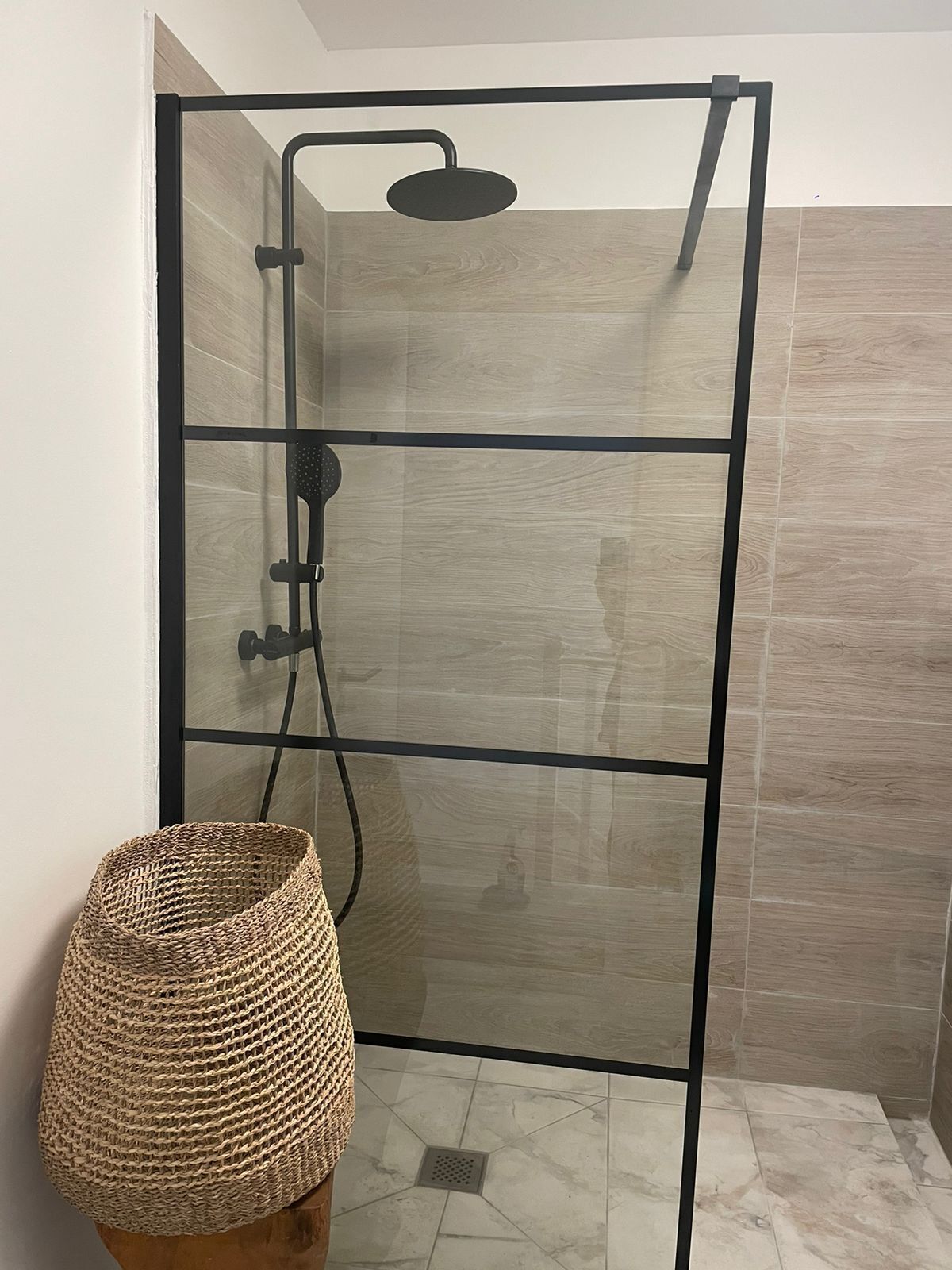 A glass-enclosed shower with a black frame, a woven basket, and a tiled floor.