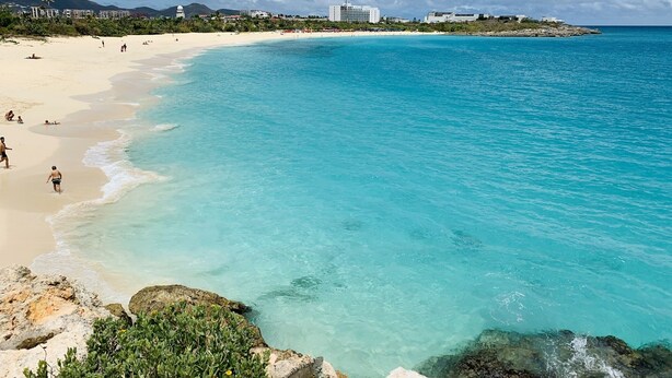 Een strand met helder turquoise water, een zandstrand en gebouwen op de achtergrond.