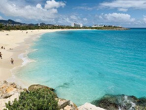 A beach with clear turquoise waters, sandy shore, and buildings in the background.