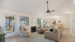 Un salon spacieux avec un grand canapé, une table basse et une cheminée. Il y a un ventilateur de plafond et une vue sur l’extérieur grâce à de grandes fenêtres.