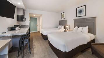 A hotel room with a kitchenette, two beds, a desk, and a chair.