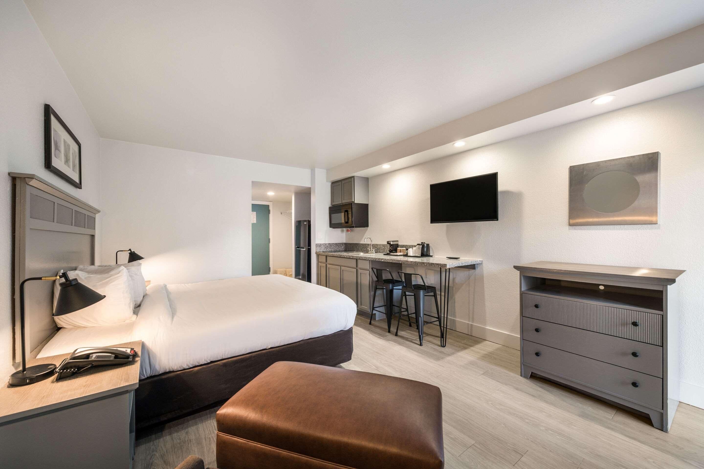A modern hotel room with a bed, desk, chair, and a kitchenette.