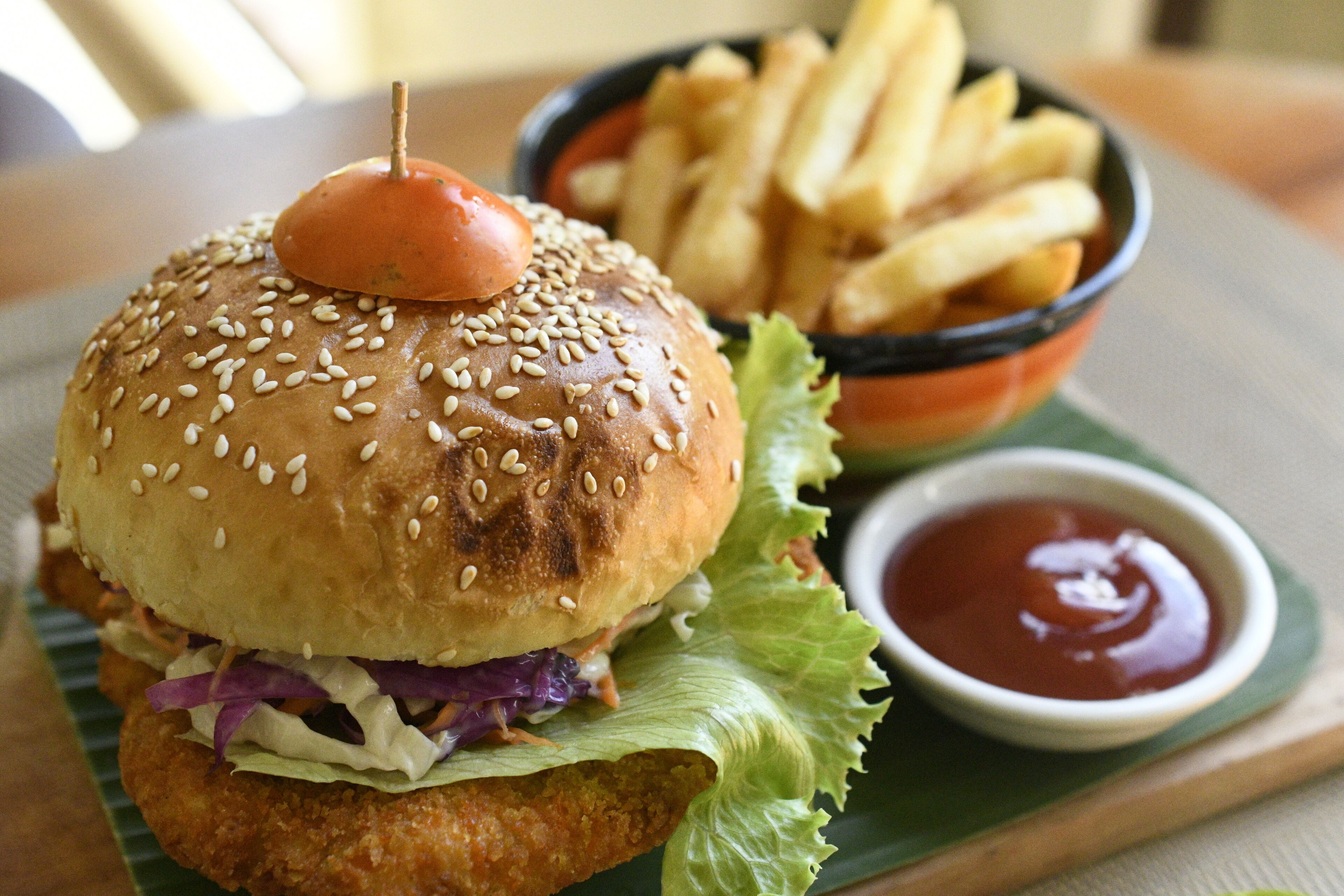 A burger with a sesame seed bun, a fried patty, lettuce, and a tomato on top, served with French fries and ketchup.