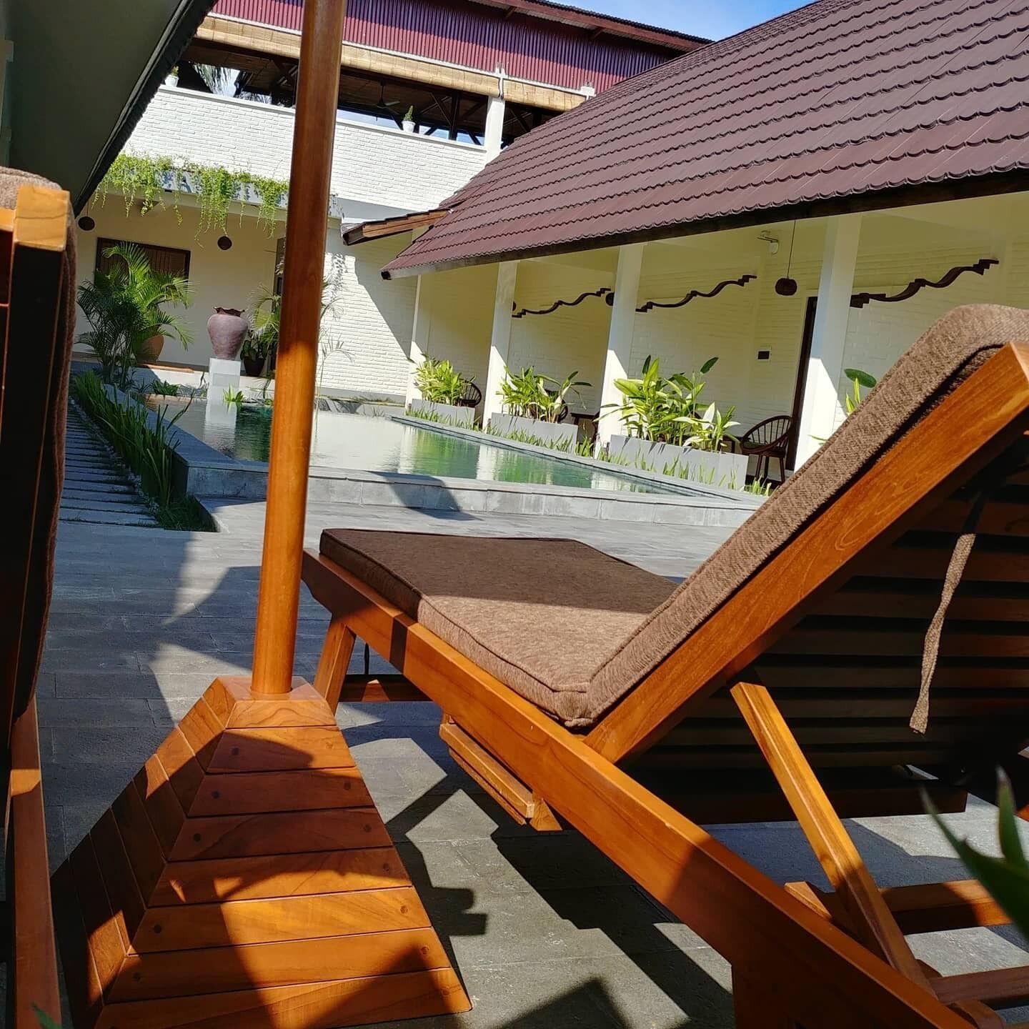 A wooden lounge chair with a cushion, a small pool, and a building with a tiled roof in the background.