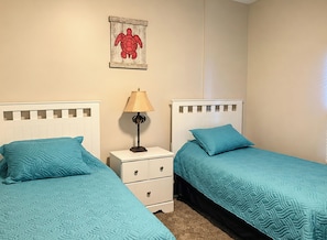 A bedroom with two beds, each with a teal bedspread, a nightstand with a lamp, and a wall-mounted picture of a turtle.
