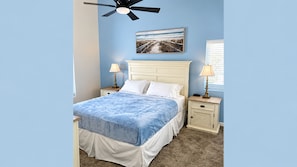 Un dormitorio con cama, dos mesitas de noche, ventilador de techo, ventana con persianas y un cuadro colgado en la pared.
