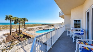 A balcony with a pool, beach view, and outdoor seating.