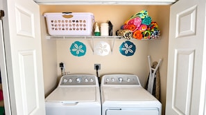 A laundry room with two front-loading washers, a shelf above with laundry baskets and folded clothes, and decorative wall stickers.