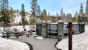 A stone fire pit with chairs, a pool, and a stone wall enclosure in a snowy outdoor area.