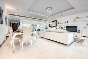 A modern living room with a white sofa, dining table, and chairs. There is a TV and a chandelier.